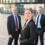 Happy beautiful business woman wearing office suit, standing outdoors and looking at camera. Male business colleagues standing behind. Business team concept